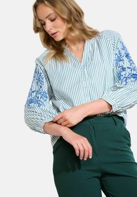 Camicia a righe blu chiaro e bianco con ricami floreali blu sulle maniche a sbuffo. Abbinata a pantaloni aderenti verde scuro.