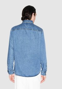 Camicia in denim blu con maniche lunghe, polsini con bottoni e orlo curvo. Presenta cuciture visibili e un dettaglio sullo yoke posteriore.