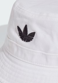 Bijela ribarska kapa izrađena od pamuka. Ima crni izvezeni logo s tri stilizirana lista s prednje strane. Struktura s teksturom i detaljima šivanja.