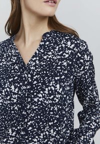 Blusa navy con motivo astratto bianco, con abbottonatura frontale e scollatura a V. Maniche lunghe con leggero arricciamento sulle spalle. Tessuto morbido.