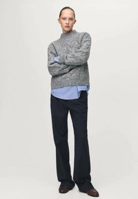 Szary dziergany sweter z wysokim kołnierzem nałożony na jasnoniebieską, pasiastą koszulę; w połączeniu z ciemnymi spodniami i brązowymi butami.