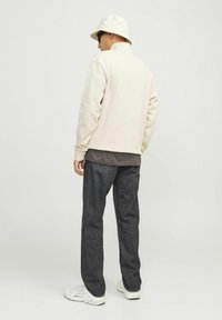 Lichtbeige fleece jack met een hoge kraag, gecombineerd met donkergrijze denim broek en witte sportschoenen. Een bucket hat en zonnebril zijn zichtbaar.