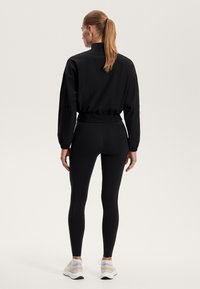 Černá lehká bunda s vysokým límcem a elastickým pasem, kombinovaná s černými legínami a bílými teniskami, předvádějící štíhlý sportovní vzhled.