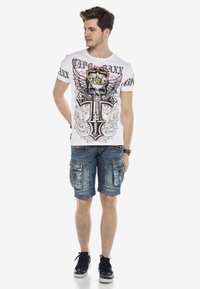 Weißes T-Shirt mit Grafik von einem Schädel, Flügeln und einer Krone; blaue Denim-Cargo-Shorts mit mehreren Taschen; schwarze Sneakers mit weißen Sohlen.