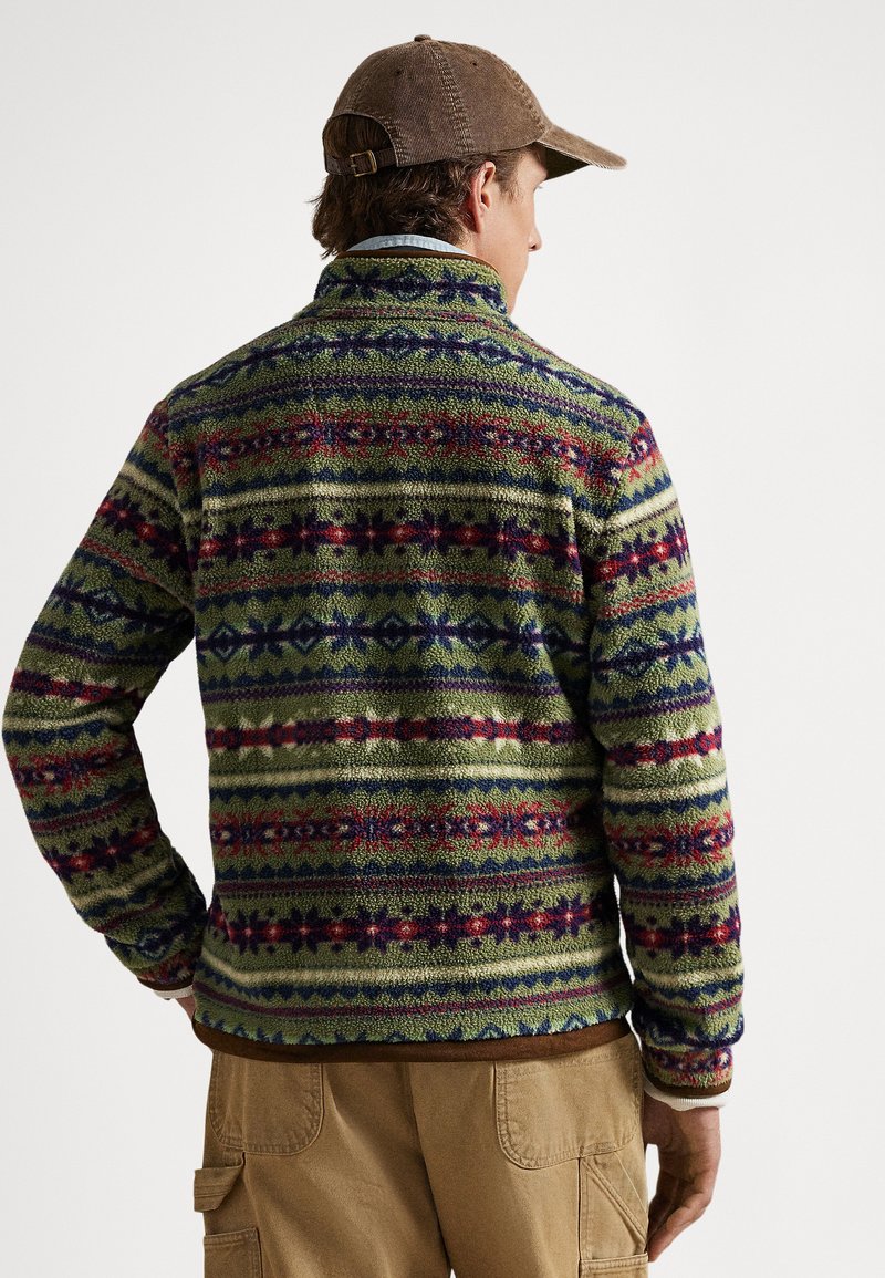 Veste en polaire présentant des motifs géométriques multicolores sur un fond vert, avec un col haut et une finition texturée. Accents marron à l'ourlet.