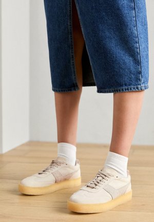 Baskets beiges avec des textures en daim et en maille, lacets blancs et semelle en caoutchouc. Associées à des chaussettes blanches et une jupe midi en denim foncé.