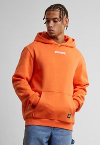 Oransje hettegenser med frontlomme og snøring i hetta. Har en hvit "STARTER" logo på brystet. Myk, teksturert stoff.