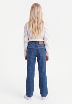 Barn med langt blondt hår, iført en langærmet, stribet trøje, blå jeans og hvide sneakers, vendt væk fra betrakteren.