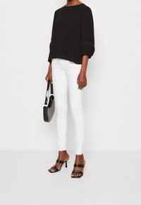 Haut noir texturé à manches larges, jean skinny blanc avec fermetures éclair à la cheville, et sandales à talons noires. Tenant un sac à main noir.