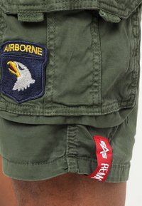 Olivgrön bomullsshorts med en blå och gul "AIRBORNE"-patch med en örn, samt en röd "REMA"-etikett i fållen. Sömnadsdetaljer synliga.