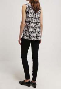 Top floreale nero senza maniche con motivi rosa e bianchi, abbinato a jeans skinny neri. Il modello indossa scarpe nere; la texture del tessuto è liscia e visibile.
