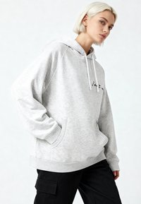 Felpa con cappuccio grigio chiaro con coulisse, tasca frontale e dettagli in scrittura. Caratterizzata da una vestibilità morbida e una texture soffice. Abbinata a pantaloni neri.