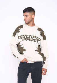 Crème trui met groene camouflagepatronen en vetgedrukte tekst "MULTIPLY APPAREL". Voorzien van geribbelde manchetten en een ronde halslijn. Gemaakt in combinatie met donkere pants.