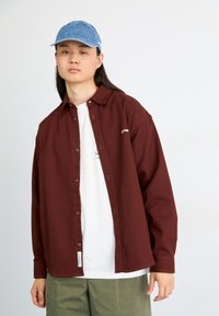 Burgundy button-up shirt made of heavy fabric, featuring a white logo detail, paired with a white t-shirt and olive green pants. Blue cap on head.