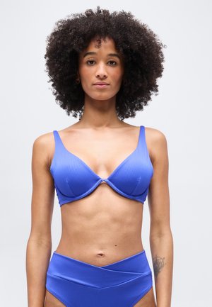 Femeie cu păr creț, purtând un top de bikini albastru cu dungi și slipuri cu talie înaltă, stând pe un fundal deschis simplu.