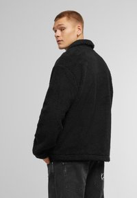 Giacca in pile nera con superficie testurizzata e morbida, colletto alto e vestibilità rilassata. Indossata con jeans scuri, mostra uno stile casual.