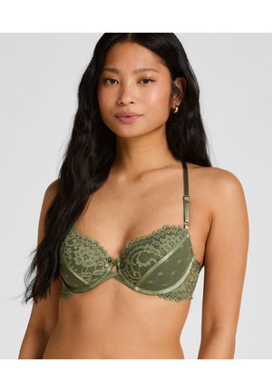 Frau mit langem schwarzen Haar, die einen olivgrünen Spitzen-BH mit verstellbaren Trägern und gezackten Rändern trägt, schaut nach vorne vor einem schlichten Hintergrund.