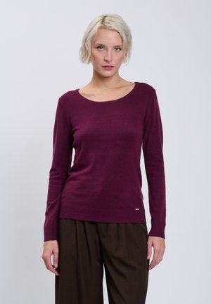 Femme aux cheveux blonds courts portant un pull violet à manches longues et un pantalon marron foncé sur un fond clair uni.