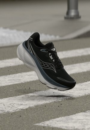 Chaussure de course noir et blanc en l'air au-dessus d'un passage piéton sur une route en asphalte gris avec de petites particules de gravier en dessous.
