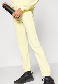 Κίτρινη φόρμα από μαλακό ύφασμα με ελαστικά μανσέτες, συνδυασμένη με μαύρα αθλητικά παπούτσια. Στο δεξί χέρι κρατιέται μια μαύρη παγούρι νερού.