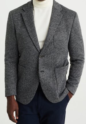 Mann trägt grauen Fischgräten-Blazer über weißem Rollkragenpullover mit dunkler Hose, eine Hand in der Tasche, vor einfarbigem Hintergrund.