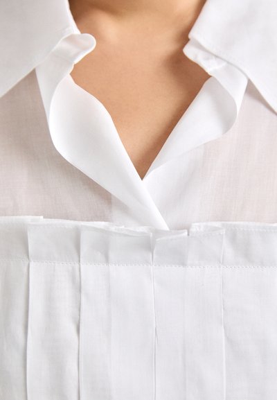 Blusa bianca con un ampio colletto, tessuto morbido e dettagli plissettati strutturati anteriori.