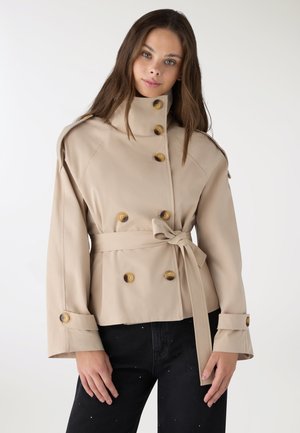 Donna che indossa una giacca beige doppiopetto con cintura e grandi bottoni, in piedi davanti a uno sfondo bianco uniforme.