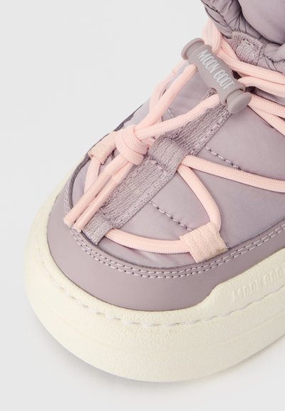 Bota roxa com parte superior sintética lisa, apresentando atacadores elásticos rosa e uma sola branca chunky. Logótipo notável na lateral.