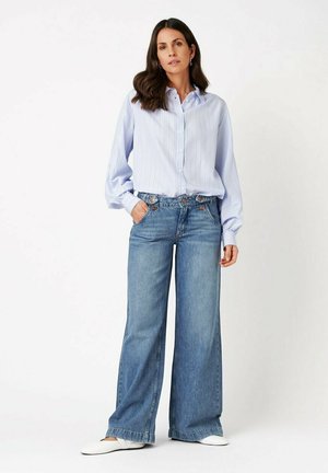 Woman stands with hands in pockets wearing light blue striped blouse, wide-leg blue jeans, and white shoes against a plain white background.