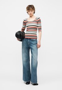 Νεαρός/ή όρθιος/α, φοράει ριγέ κοντομάνικη μπλούζα, μπλε τζιν παντελόνι με φαρδιά πόδια, μαύρα παπούτσια και κρατά μια μαύρη δερμάτινη τσάντα.