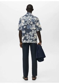 Cămașă scurtă cu mâneci, de culoare albastră, cu motive florale și modele albe, asortată cu pantaloni negri. O jachetă bleumarin este ținută într-o mână.