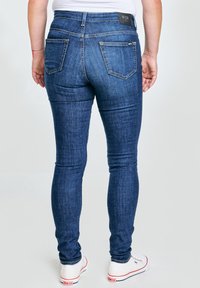 Osoba v přiléhavých modrých džínách a bílých teniskách, zobrazena zezadu na jednoduchém světlém pozadí.