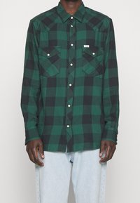 Zielona i czarna kraciasta koszula flanelowa z długimi rękawami, dwoma kieszeniami na piersi i zatrzaskami; materiał wydaje się miękki i teksturowany.
