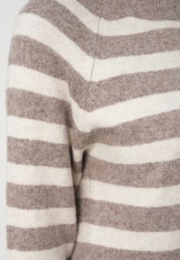Dryžuotas megztinis iš minkštos smėlio spalvos ir šviesiai rudos vilnos. Turi plačias horizontalias juostas, apvalų iškirptę ir laisvą siluetą. Tekstūruota megzta medžiaga.