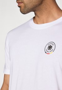 Balta kokvilnas T-krekls ar apaļu kakla izgriezumu un īsām piedurknēm, ar apaļu logotipu, kurā attēlots melns ērglis un sarkani-dzelteni akcenti.