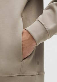 Beige sweatshirt met een zachte textuur, een kangoeroezak en een ribgebreide manchette. Klein logo detail zichtbaar aan de onderkant van voren.