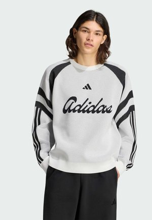 Junge Person mit mittellangen dunklen Haaren, die einen weißen Adidas-Pullover mit schwarzen Streifen und schwarze Hose trägt, mit Händen in den Taschen.