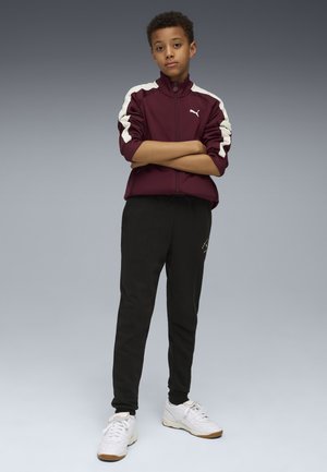 Maroon zip-up jacket with white accents, paired with black pants. Model wears white sneakers. Background fades from gray to dark.