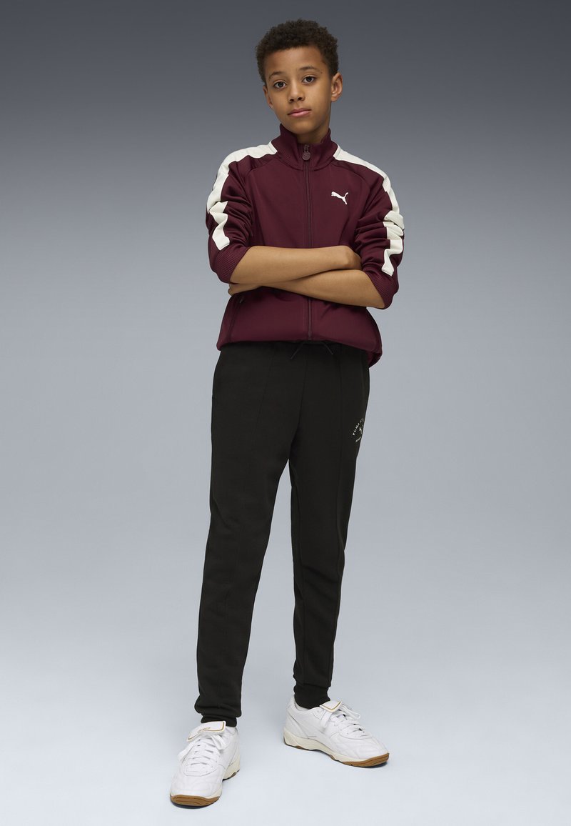 Bordeauxfärgad zip-up jacka med vita accenter, kombinerad med svarta byxor. Modellen bär vita sneakers. Bakgrunden övergår från grått till mörkt.