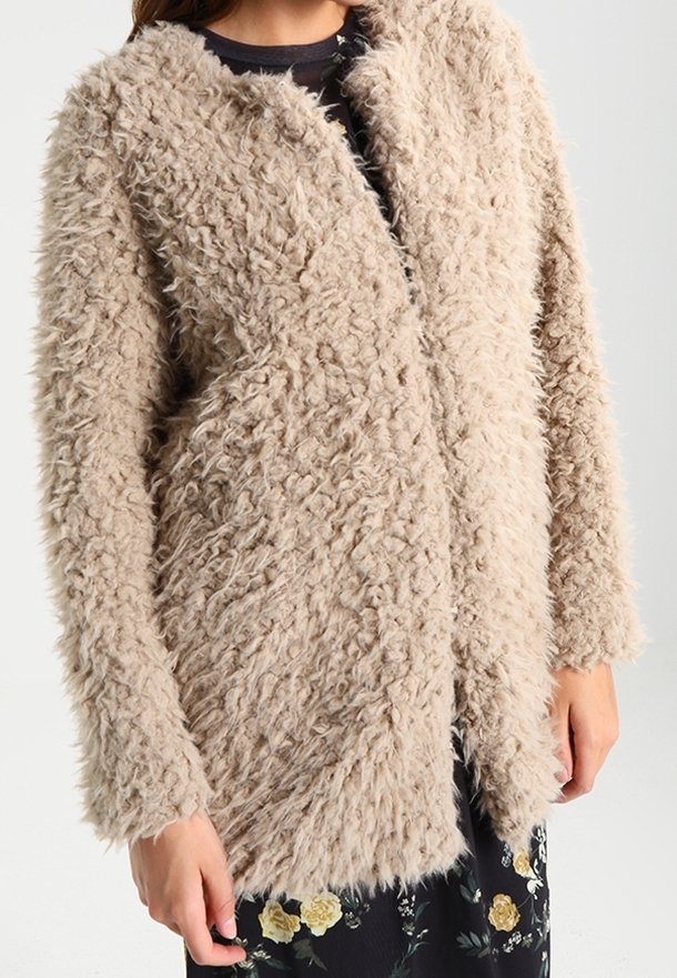 Femme portant une veste longue à manches longues, texturée et moelleuse de couleur beige, par-dessus une robe fleurie noire, les bras détendus le long du corps.