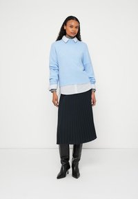 Svetlomodrý nadrozmerný sveter cez bielu košeľu s golierom, v kombinácii s plisovanou čiernou sukňou a čiernymi vysokými čižmami po kolená. Jednoduché pozadie.