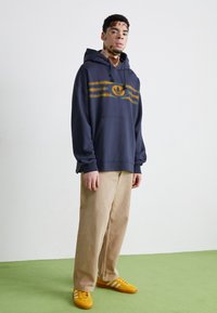 Tumed sinine kapuutsiga sviiter kollaste triipudega ja logoga, koos beežide laia säärega pükstega ning kollaste vihmavarjuga valgete triipudega. Sile kangatekstuur.