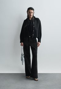 Woman standing against a plain wall wearing black jacket, black pants, black sandals, and holding a small silver handbag.