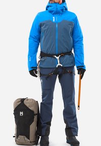 Blau-graue wasserdichte Jacke mit Kapuze, kombiniert mit grauen Hosen. Beinhaltet ein Geschirr, Handschuhe und eine orangefarbene Eisäxte. Schwarzer und brauner Rucksack.
