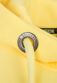 Gelbes Gewebe mit einer glatten Textur zeigt ein Metallösen mit der Gravur "SPITZBUB" und einem geflochtenen Kordelzug, der hindurchgezogen ist.