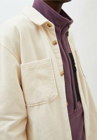 Crèmekleurige corduroy shirtjacket met gouden knopen, enkele borstzak; gelaagd over een paarse fleece top met rits. Gestructureerde stof zichtbaar.