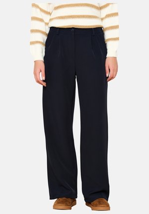 Žena v černých širokých kalhotách, bílo-béžovém pruhovaném svetru a hnědých nazouvacích botách, stojící před jednobarevným pozadím.