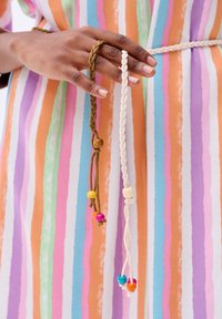 Dwie plecione sznurki, jeden oliwkowo-zielony z kolorowymi koralikami, a drugi biały z drewnianą koraliką, trzymane w dłoni na tle w paski w różnych kolorach.