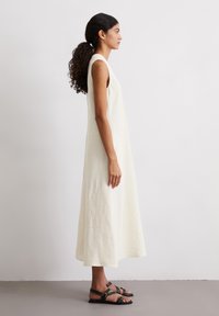 Weißes, ärmelloses Leinenkleid mit fließender Silhouette. Verfügt über einen runden Ausschnitt und reicht bis zur Mitte der Wade. Kombiniert mit schwarzen Riemchensandalen.