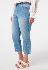 Jean en denim bleu clair, de coupe courte, orné de perles blanches le long de l'ourlet. Assorti à des sandales à talons tressées beiges.